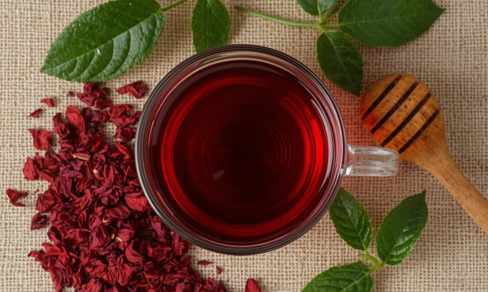 Top view of a glass cup filled with red hibiscus tea surrounded by dried petals, green leaves, and a wooden honey spoon on linen fabric
