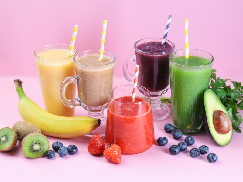 Five colorful smoothies displayed on a pink background, including fruit-based and green smoothies, with fresh ingredients such as banana, strawberries, blueberries, kiwi, avocado, and cucumber
